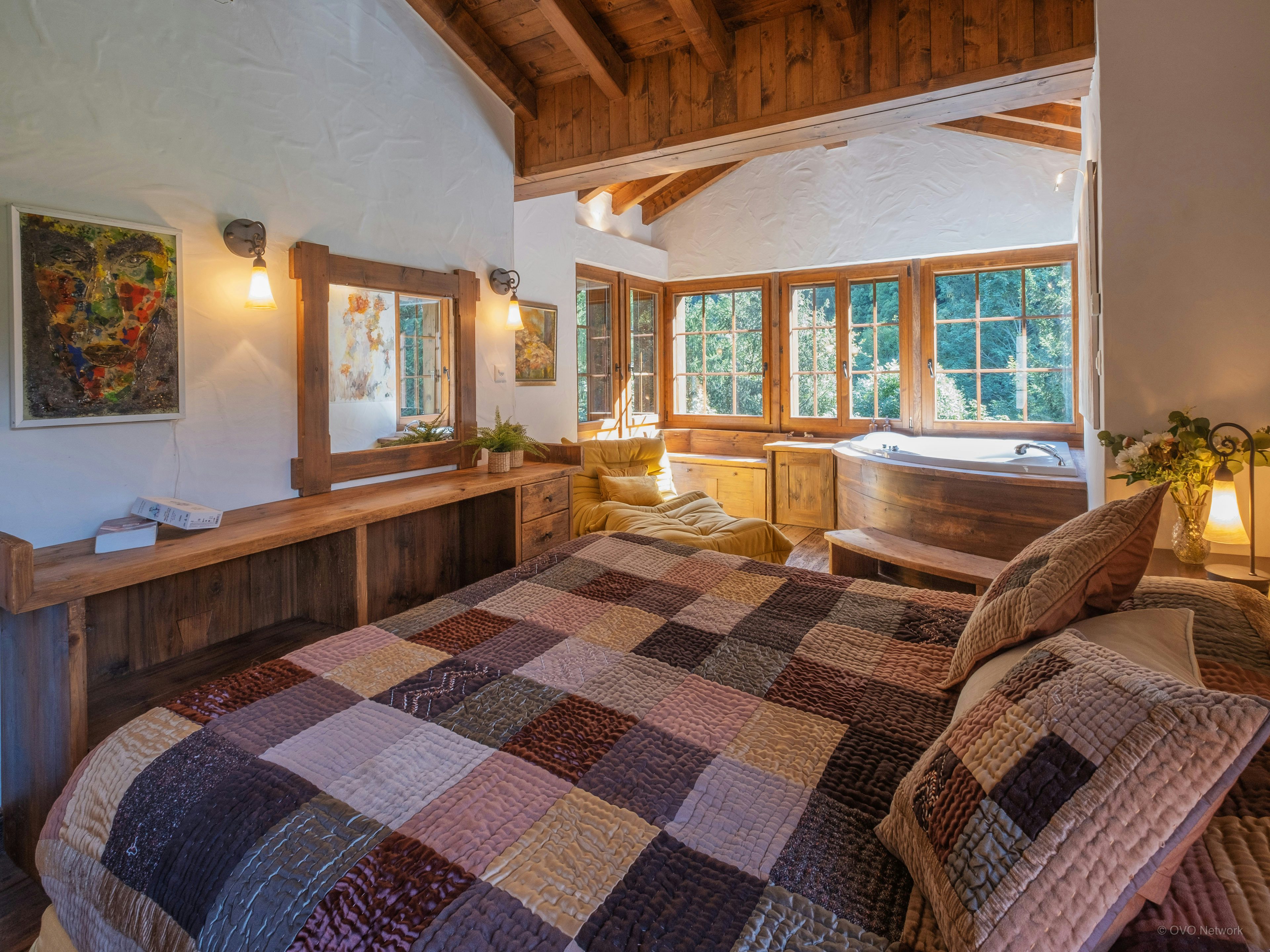 L'une des chambres du chalet avec un grand lit et une baignoire d'angle, donnant vue sur la nature environnante grâce à la présence de grandes fenêtres.